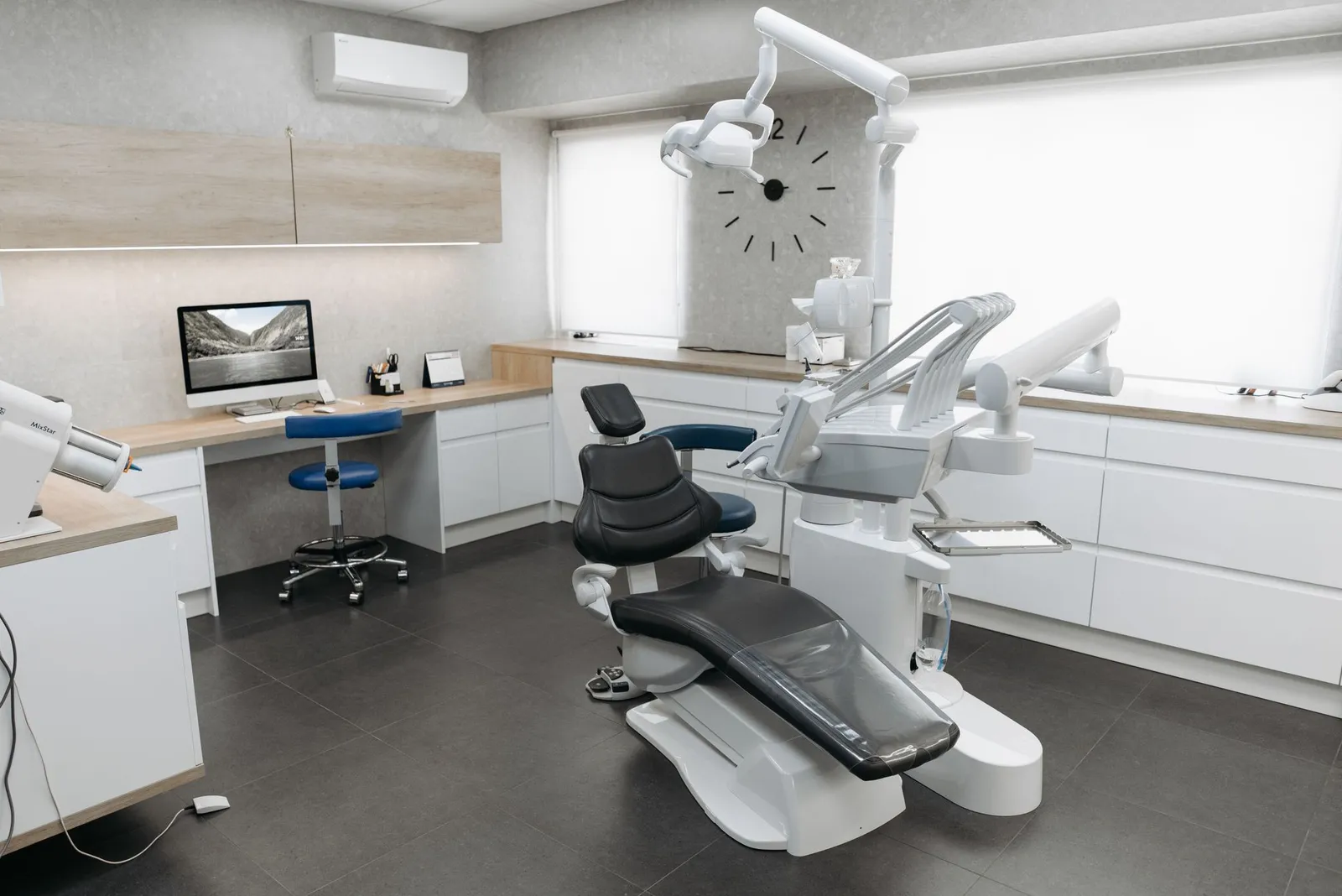 A bright, modern dental treatment room with a dental chair, overhead light, wood-toned cabinetry, and large windows