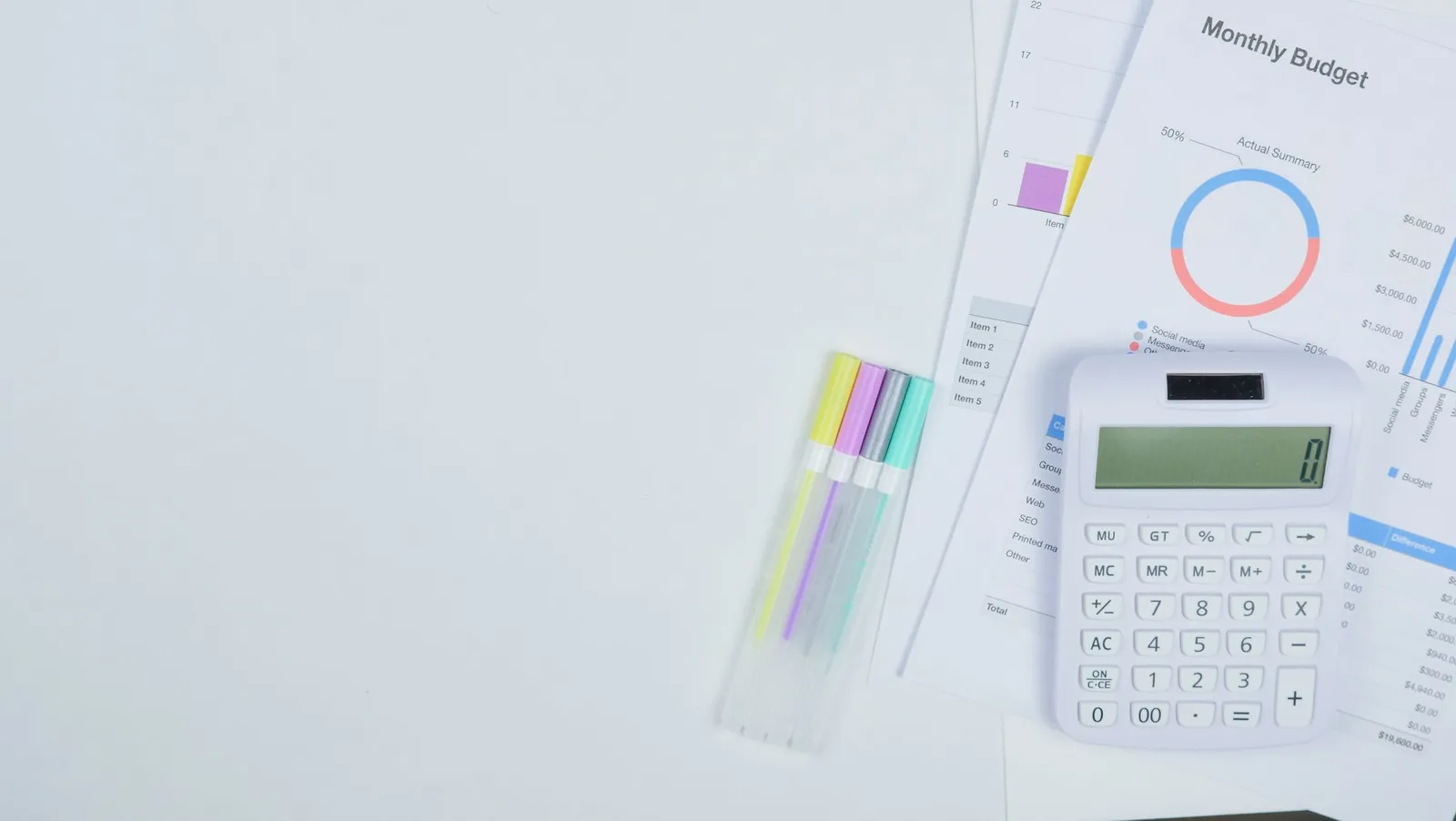 A calculator sits on a desk next to a printed monthly budget report with colorful pens beside it