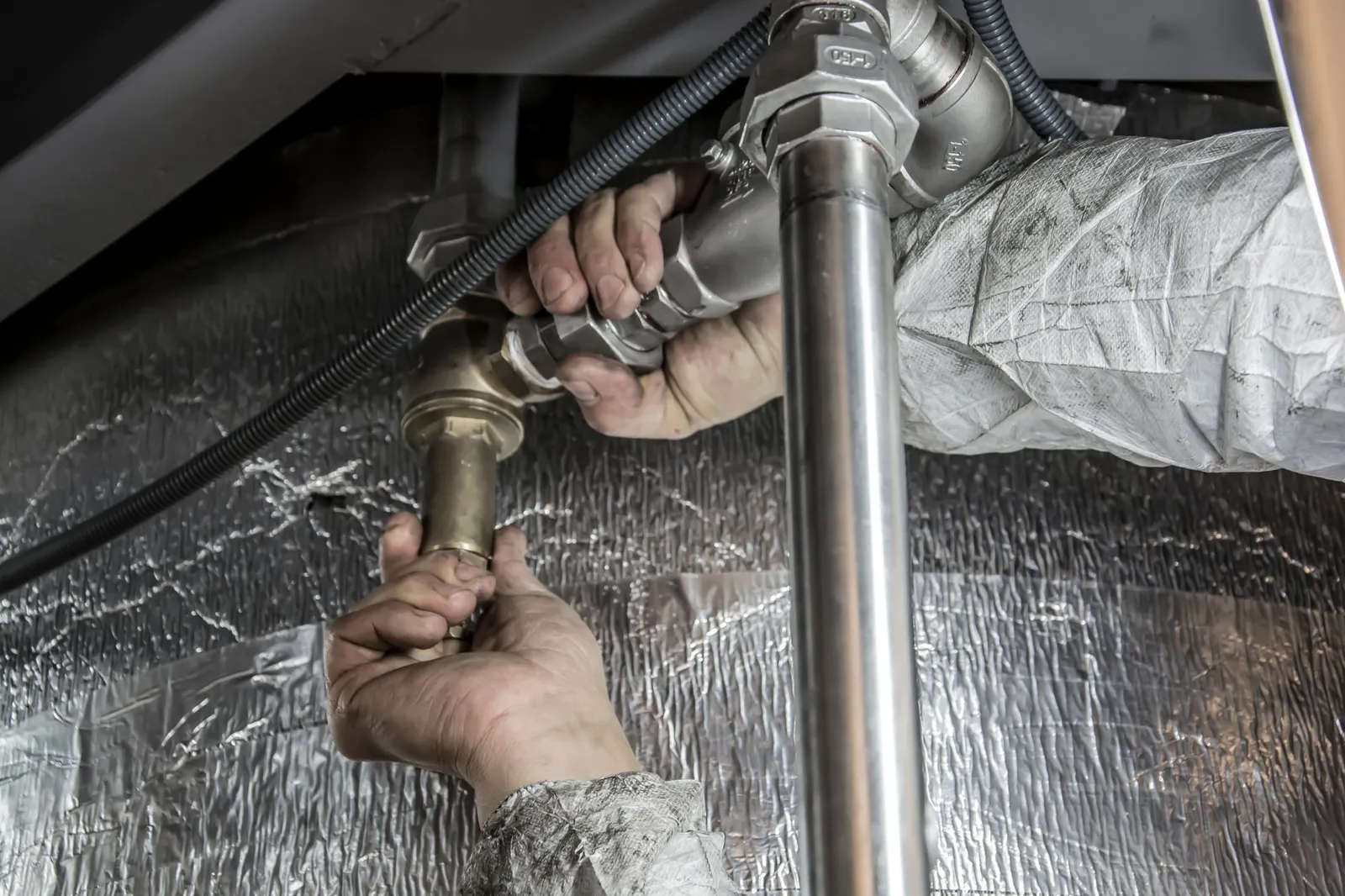A plumber's hands working with a wrench on steel pipes under a sink