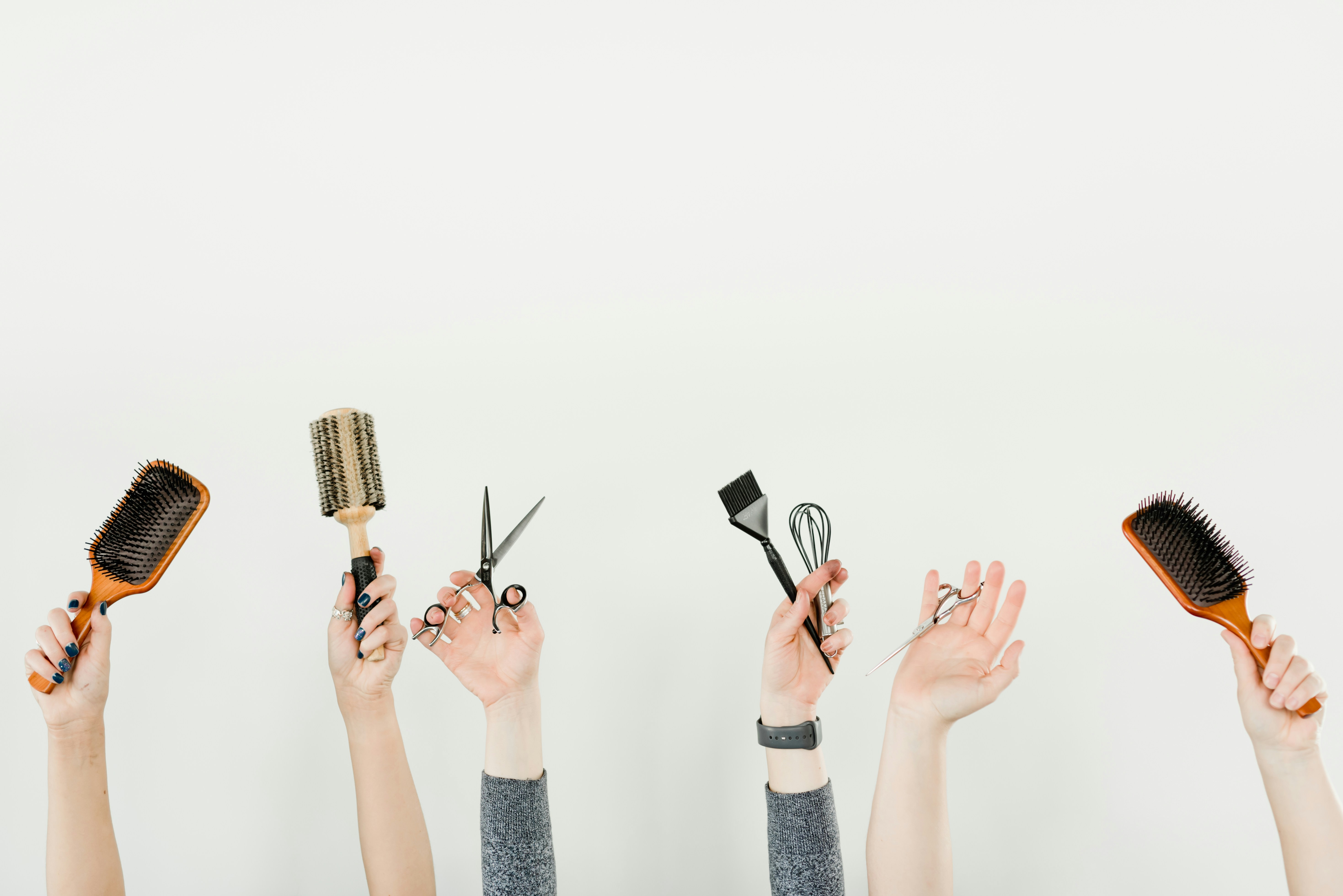 Hands holding hairbrushes, scissors, and a tinting brush against a white background