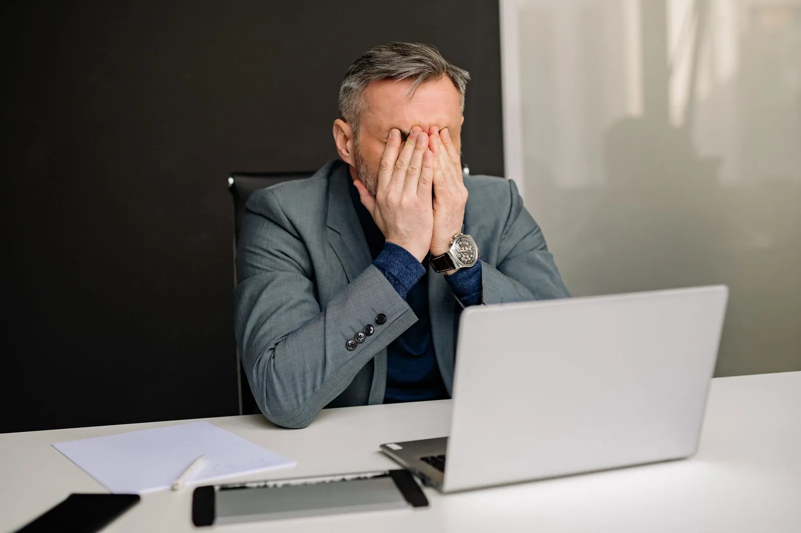 A frustrated business owner in a gray suit sits with his head in his hands at a desk in front of an open laptop