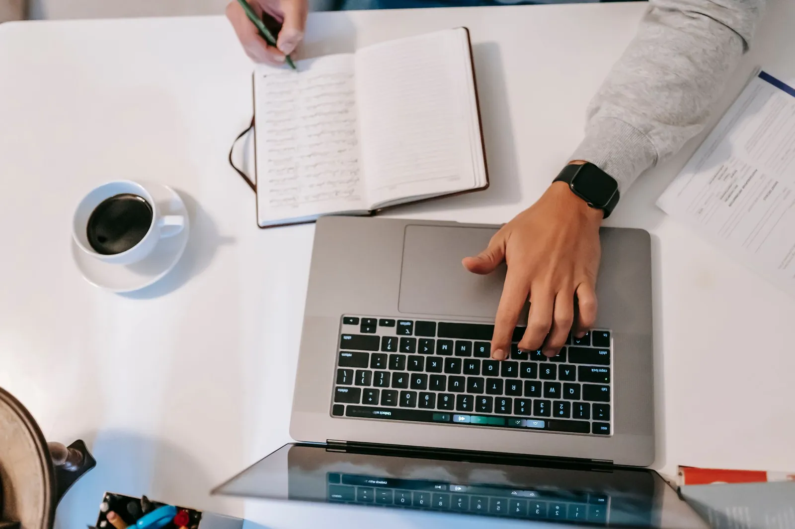 Overhead view of a laptop and a handwritten notebook on a desk, with a cup of coffee beside them